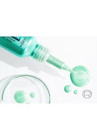 Gel exfoliant turquoise dans un tube en plastique avec un embout de précision, accompagné de gouttes circulaires de produit sur un fond blanc.
