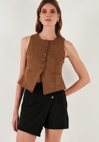 Barna szarvasbőr ujjatlan mellény, testhezálló fazonban, gombos elülső részel és két oldalsó zsebbel, fekete szarvasbőr sorttal párosítva.