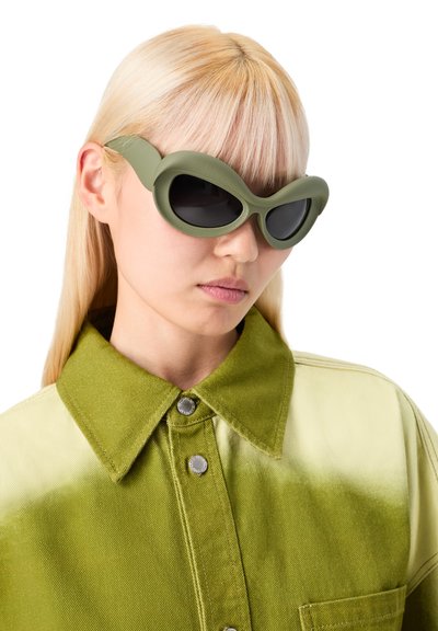 Zelené sluneční brýle ve tvaru kočičích očí s černými skly; texturovaný rám; nošeny s dvoubarevným zeleným denimovým tričkem s velkým límcem a detailem knoflíků.