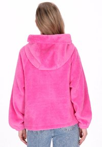 Vaaleanpunainen fleece-takki hupulla, jossa pehmeä pinta, väljä istuvuus ja laskostetut olkapäät. Takakuvassa on yksinkertainen malli ilman koristeluja.