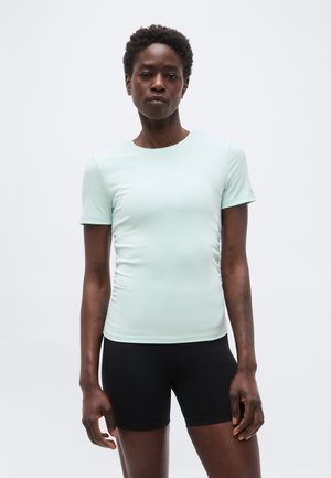 Γυναίκα με κοντά σγουρά μαλλιά που φοράει ανοιχτό μέντα πράσινο κοντομάνικο τοπ και μαύρο εφαρμοστό σορτς, στέκεται μπροστά από ένα απλό λευκό φόντο.