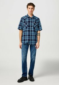 Camicia a maniche corte a quadri in tonalità blu con sfondo scuro, dotata di due tasche sul petto. Abbinata a jeans blu e scarpe nere.