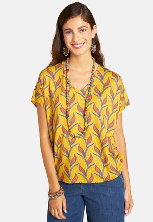 Woman smiling wearing a yellow patterned blouse with orange and purple leaves, blue jeans, and colorful beaded necklace, earrings, and bracelet.