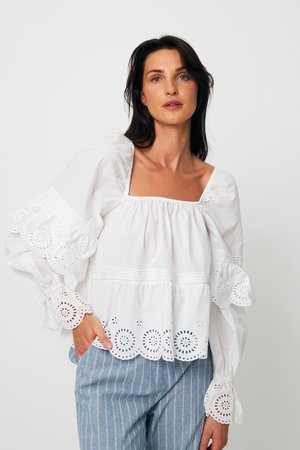 Γυναίκα που φοράει λευκό πουκάμισο με μακριά μανίκια, διακοσμημένο με δαντέλα eyelet και φουσκωτά μανίκια, συνδυασμένο με μπλε ριγέ παντελόνια, στέκεται μπροστά σε απλό φόντο.