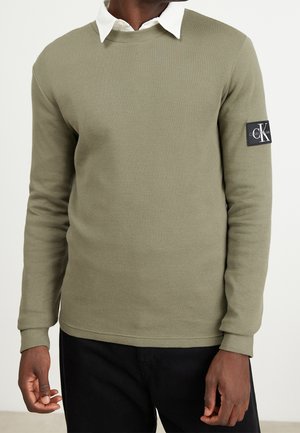 Homme portant un pull texturé vert olive par-dessus une chemise blanche à col, avec un pantalon noir et un patch logo Calvin Klein sur la manche gauche.