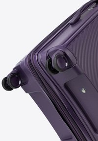Valise rigide violette avec une surface texturée, double roulettes et fermeture éclair. Présente des bords arrondis et une construction robuste.