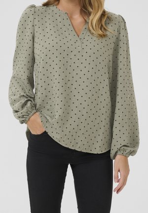 Kvinde iført lyse olivengrøn bluse med sorte polkaprikker, lange pufærmer, V-hals og sorte bukser, hånd i lommen.