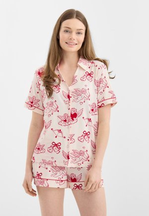 Jonge vrouw die bijpassende lichtroze pyjama draagt met rode Cupido- en hartafdrukken, staande tegen een effen witte achtergrond.