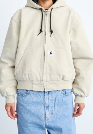 Person iført beige lynlåstrøje med hætte og sorte snore samt lyseblå jeans, der står mod en ensfarvet baggrund.