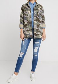 Chaqueta con patrón de camuflaje y dos bolsillos frontales, llevada sobre una camisa de mezclilla azul, combinada con jeans azules desgastados y zapatillas blancas.