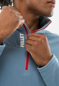 Pull en polaire bleu texturé avec un col zippé, présentant des accents rouges et un logo "MILLET" blanc sur le devant.