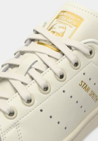 Valge nahkne tenniseki, millel on perforeritud küljed, lamedad beežid paelad ja kuldne logo silt keele peal. Omab minimalistikku disaini ja ümarat kuju.