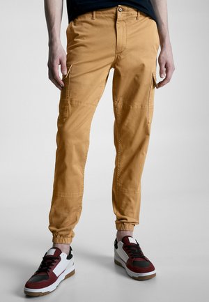 Pantalons cargo en coton jaune moutarde, avec des poches latérales, des poignets élastiques et une coupe décontractée. Associés à des baskets noires et rouges.