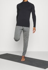 Person balancing on one barefoot on a yoga mat while holding the other foot behind, wearing a black top and gray pants.