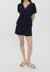Námořnická modrá, krátký rukáv, top ve stylu převázání s peplum designem, v kombinaci s odpovídajícími šortkami; oboje má texturovanou látku s otvorovými vzory. Sandály doplňují celkový vzhled.