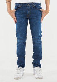 Jeans in denim blu con una vestibilità slim, caratterizzati da cuciture arancioni, un bottone frontale e cinque tasche. Indossati con scarpe da ginnastica bianche.