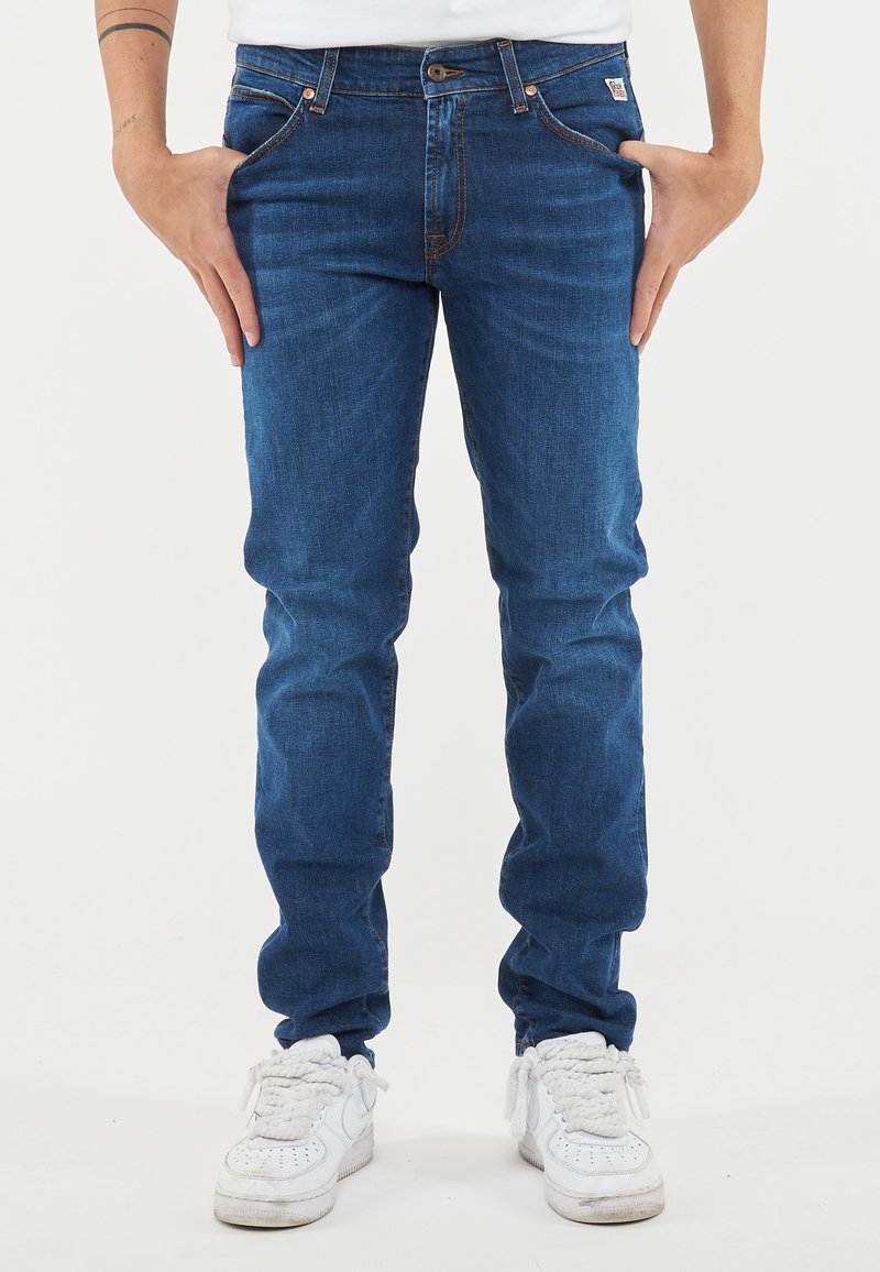 Jeans in denim blu con una vestibilità slim, caratterizzati da cuciture arancioni, un bottone frontale e cinque tasche. Indossati con scarpe da ginnastica bianche.