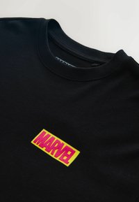 Mustast puuvillast T-särk ümar kaelusega, esiküljel silmatorkava kollase ja roosa "MARVEL" logo plaastriga. Sile tekstuur.