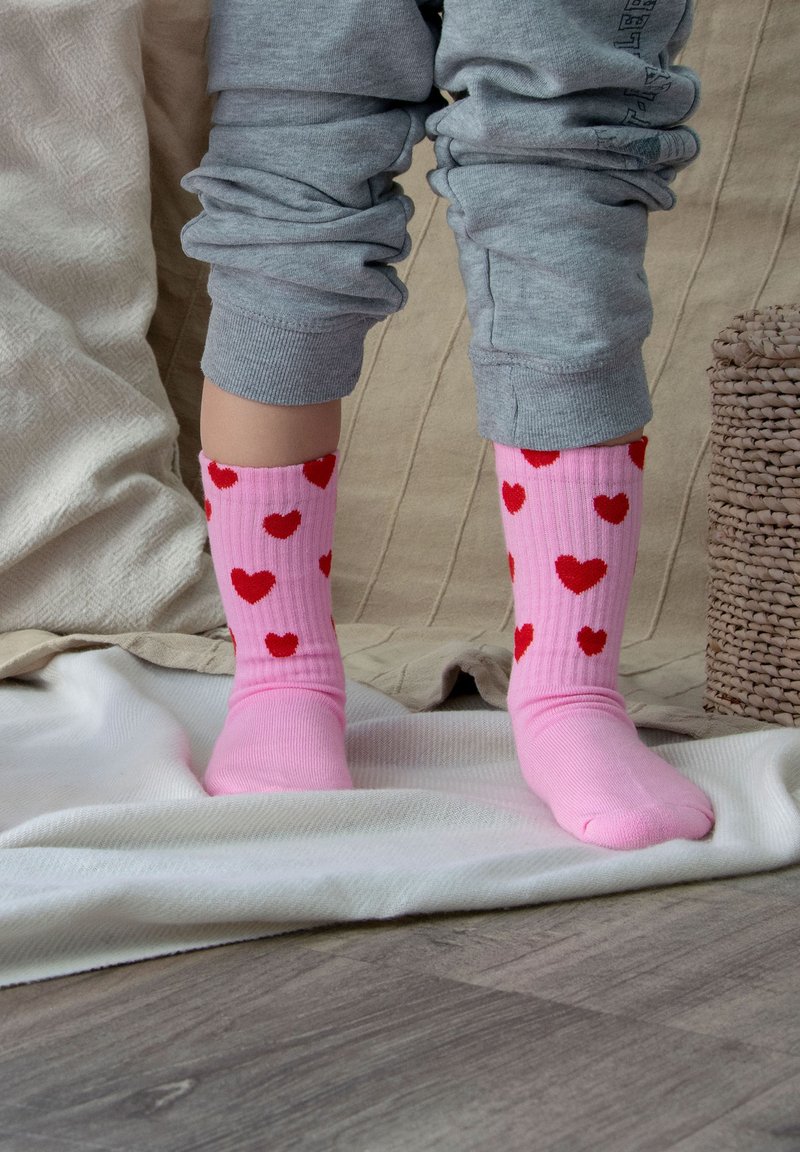 Chaussettes roses avec des motifs de cœurs rouges, design côtelé, ajustées en haut. Portées avec un pantalon de jogging gris, debout sur une couverture de couleur claire.