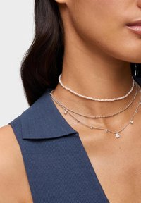 Warstwowe naszyjniki złożone z srebrnego łańcuszka z małymi koralikami, różowego naszyjnika choker z koralików oraz zawieszki dekoracyjnej, noszone na tle niebieskiej tkaniny.