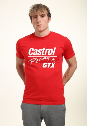 Ung mand iført en rød Castrol Racing GTX t-shirt og grå bukser, stående med hænderne bag ryggen mod en ensfarvet baggrund.