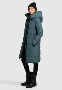 Polstret teal lang frakke med høj krave, sidelommer med lynlås og lynlås lukning. Bæret over sorte leggings og knæhøje læderstøvler.