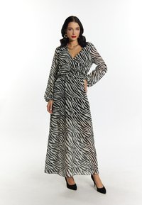 Langes Kleid mit Zebra-Print aus transparentem Stoff im Wickeldesign, akzentuiert durch eine taillierte Taille und lange Ärmel. Schwarze, hochhackige Schuhe vervollständigen den Look.