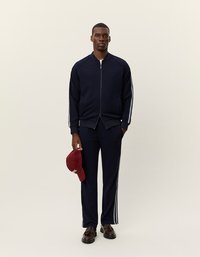 Ναυτικό αθλητικό κοστούμι με λευκές πλευρικές ρίγες, ζακέτα με φερμουάρ και παντελόνια με ελαστική μέση. Κλασικό καπέλο με επίπεδη γείσο σε μπορντό κρατημένο στο χέρι.