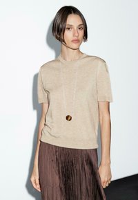 Una donna sta in piedi indossando un top lavorato a maglia beige e una gonna plissettata marrone, con una lunga collana d'argento con un pendente rotondo, su uno sfondo semplice.