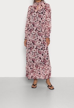 Žena v dlouhých rukávech, růžových a vínových květinových šatech až na kotníky s volánkovým límcem a černými sandály na podpatku.