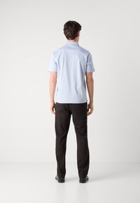 Camisa de manga curta azul clara com colarinho, combinada com calças pretas. A camisa apresenta uma textura suave e pequenas fendas laterais perto da bainha.