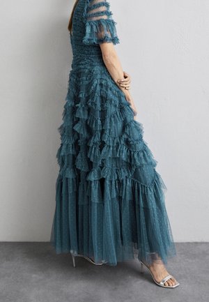 Vrouw in een tealkleurige jurk met gelaagde ruches van tule en doorschijnende mouwen met stippen, staand tegen een eenvoudige grijze muur, met zilveren sandalen met hoge hakken.