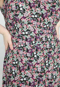 Gros plan d'une personne portant une robe à fleurs avec des fleurs vertes, roses, blanches et violettes, les mains posées sur les hanches.