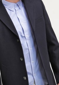 Marinblå ullmixkappa med en ensam bröstficka, stora knappar och en ljusblå skjorta med krage under.