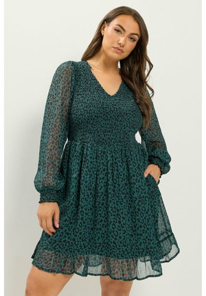 Femme portant une robe verte à imprimé léopard avec un corsage smocké, de longues manches transparentes et un ourlet volanté, debout devant un fond blanc uni.