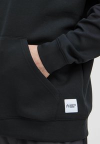 Svart sweatshirt med en framficka, ribbade muddkanter och en liten vit etikett med "NORTH BEND"-logotyp broderad på fållen.