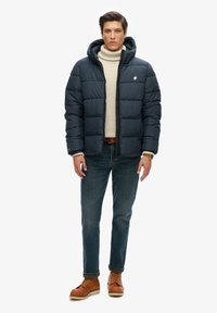 Marineblå puffjakke med hætte, der har kviltede paneler og et logo. Bæres over en beige turtleneck, parret med mørke jeans og brune støvler.