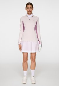 Kvinna står klädd i en ljusrosa golfoutfit med långärmad zip-up-tröja, plisserad kjol, vit handske på vänster hand och vita sneakers.