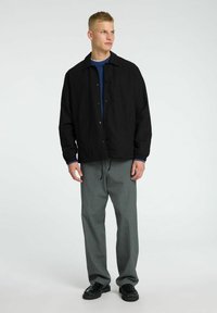 Giacca leggera nera con chiusura a bottoni, polsini elastici, abbinata a pantaloni grigi plissettati e scarpe nere. Design semplice e rilassato.