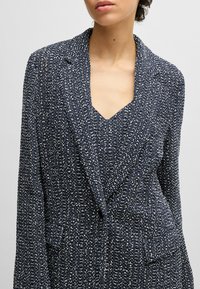 Navy textured blazer with a v-neckline, long sleeves, and a single front pocket, featuring a speckled white pattern throughout.