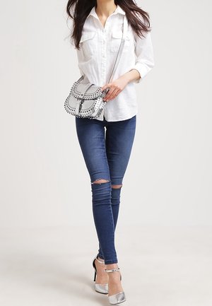 Donna che indossa una camicia bianca abbottonata, jeans skinny strappati, tacchi alti argento e porta una borsa a tracolla argento con cuciture nere.