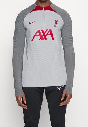Hombre vistiendo un top de entrenamiento de manga larga gris del Liverpool FC con logo rojo de AXA, cremallera hasta medio pecho, y pantalones negros de Nike con logo swoosh blanco.