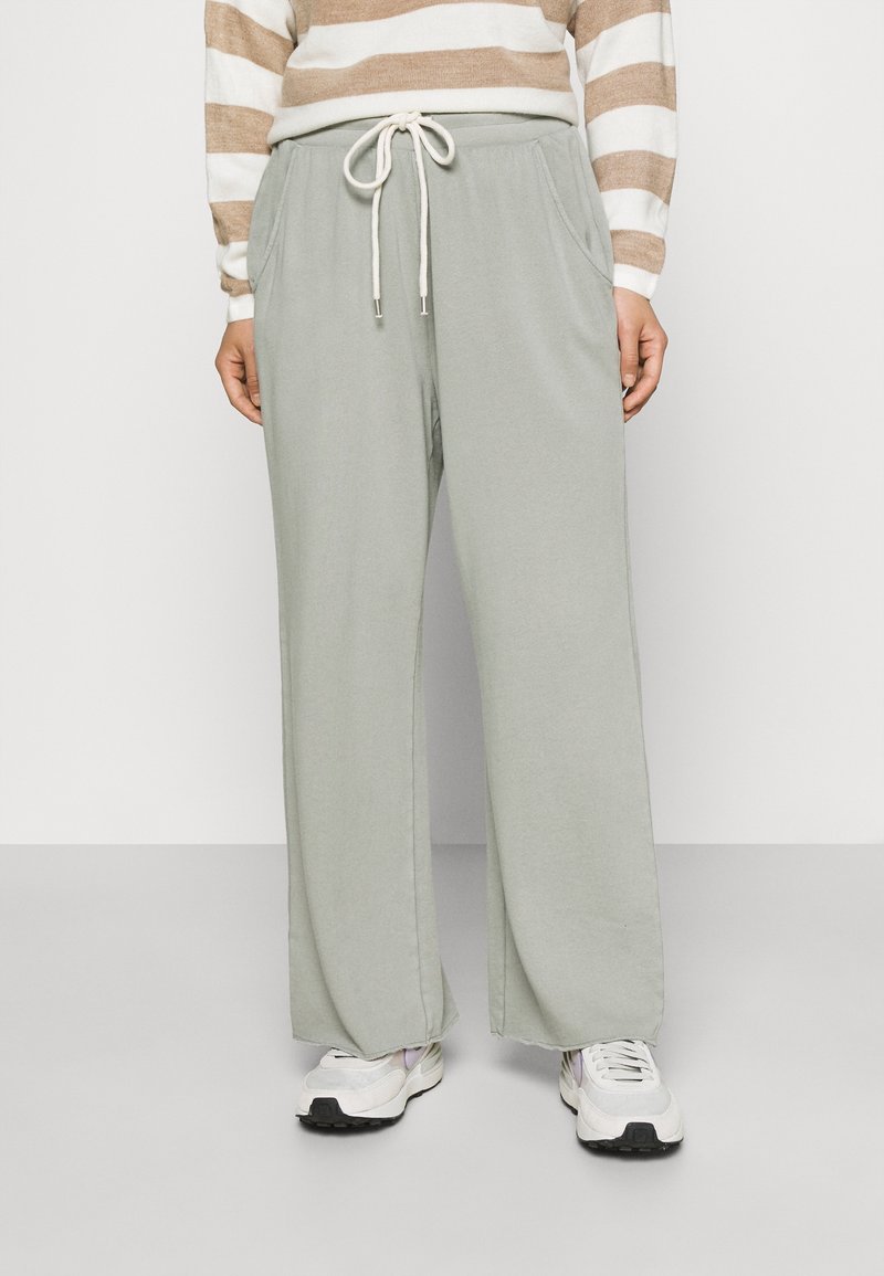 Personne portant un pantalon large gris clair avec taille à cordon, un haut à manches longues beige et blanc rayé, et des baskets blanches.