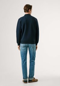 Mornarska dukserica s okruglim izrezom, rebrastim manžetama i donjim dijelom, u kombinaciji s svijetloplavenim trapericama i smeškim cipelama, prikazana straga.