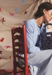 Egy világoskék kardigánban és szoknyában ülő nő piros fa széken, kezében tengerészkék virágmintás táskával, csillagmintás háttér előtt.