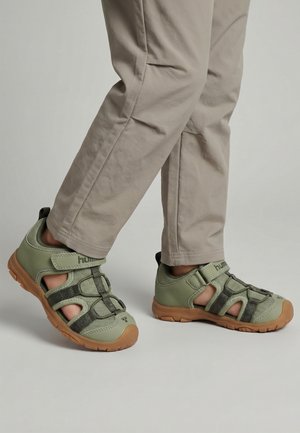 Kind trägt grüne und braune geschlossene Sandalen mit Klettverschlüssen und Mesh-Details, kombiniert mit beigen Hosen vor einem einfarbigen Hintergrund.