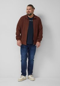 Brązowa teksturowana kurtka na zamek, na czarnej koszulce, w połączeniu z niebieskimi dżinsami i beżowymi trampkami. Model stoi na neutralnym tle.