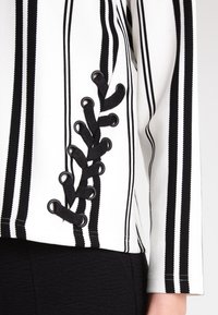 Svart och vit randig topp i ett strukturerat tyg med snörningsdetalj på sidan och öglor, som kontrasterar mot den enfärgade svarta basen.