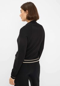 Zwarte cardigan gemaakt van zacht materiaal, met een getailleerd ontwerp, een kleurcontrasterende geribbelde tailleband en lange mouwen met subtiele accenten.