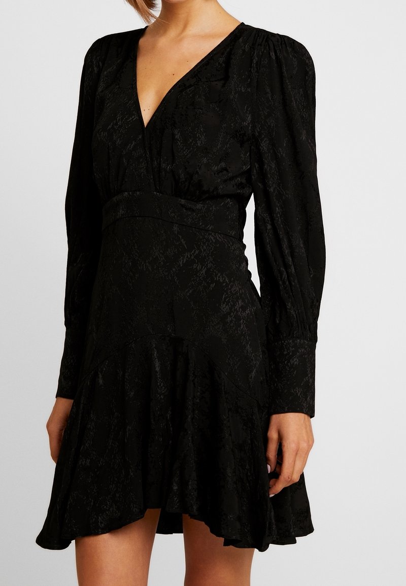 Robe noire à manches longues, col en V profond, tissu texturé, et jupe évasée avec une couture au niveau de la taille et de l'ourlet.